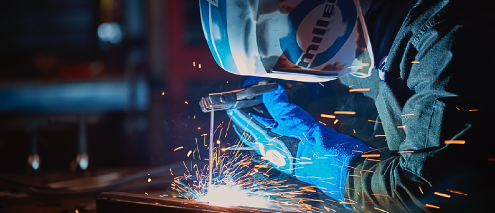 Worker wearing blue gloves and protective helmet while welding metal causing bright sparks in workshop environment